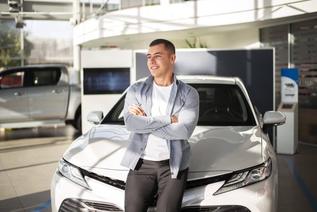 Homem sorridente de braços cruzados, encostado em um carro branco em uma concessionária, com outro veículo ao fundo.