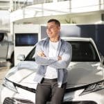 Homem sorridente de braços cruzados, encostado em um carro branco em uma concessionária, com outro veículo ao fundo.