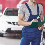 Mecânico realizando vistoria automotiva, segurando uma prancheta e uma chave inglesa, enquanto inspeciona um veículo.