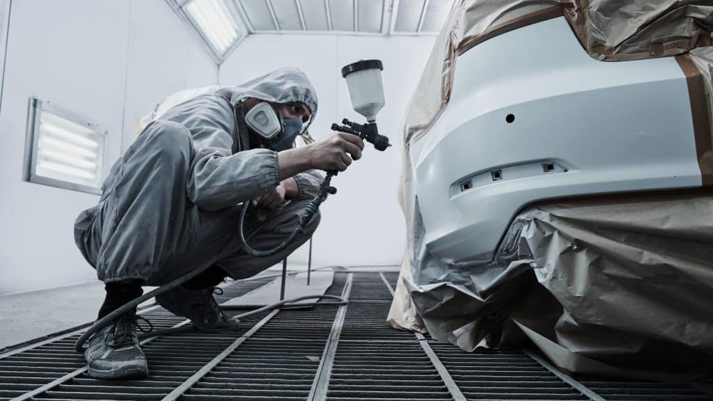 Profissional usando roupa de proteção e máscara realiza o retoque de pintura automotiva em um para-choque de carro, utilizando uma pistola de pintura em uma cabine preparada para esse tipo de serviço.