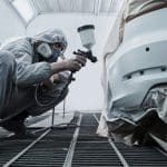 Profissional usando roupa de proteção e máscara realiza o retoque de pintura automotiva em um para-choque de carro, utilizando uma pistola de pintura em uma cabine preparada para esse tipo de serviço.