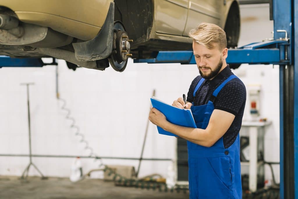 Um mecânico vestido com macacão azul e segurando uma prancheta está realizando uma vistoria em um carro elevado em uma plataforma, em uma oficina automotiva.