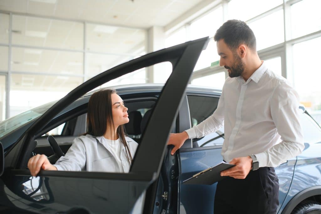 Vendedora apresentando um carro seminovo para uma cliente dentro de uma concessionária iluminada, destacando a interação profissional e o veículo em exibição.