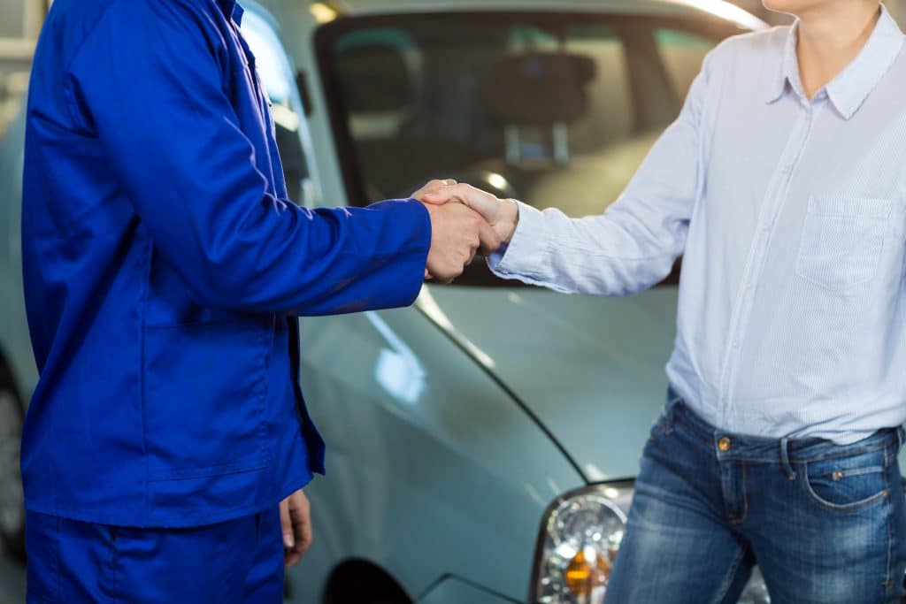 Pessoa vestindo uniforme azul apertando as mãos de um cliente em frente a um carro, simbolizando a finalização de um acordo ou compra de veículo usado.
