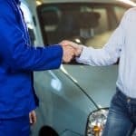Pessoa vestindo uniforme azul apertando as mãos de um cliente em frente a um carro, simbolizando a finalização de um acordo ou compra de veículo usado.