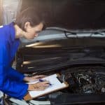 Mecânica realizando vistoria veicular, anotando informações em uma prancheta enquanto inspeciona o motor de um carro com o capô aberto, em um ambiente iluminado de oficina.