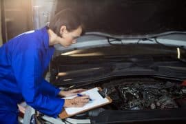 Mecânica realizando vistoria veicular, anotando informações em uma prancheta enquanto inspeciona o motor de um carro com o capô aberto, em um ambiente iluminado de oficina.