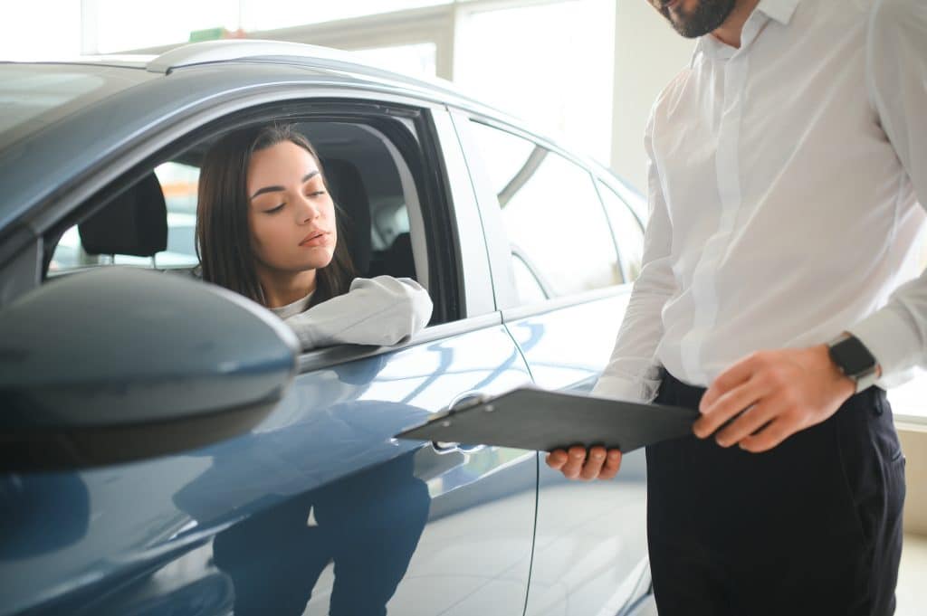 Uma mulher dentro de um carro azul observa atentamente um homem ao lado do veículo, segurando uma prancheta. A cena sugere uma negociação, avaliação ou entrega de um automóvel, possivelmente em um contexto de locação, compra ou venda.