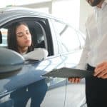 Uma mulher dentro de um carro azul observa atentamente um homem ao lado do veículo, segurando uma prancheta. A cena sugere uma negociação, avaliação ou entrega de um automóvel, possivelmente em um contexto de locação, compra ou venda.