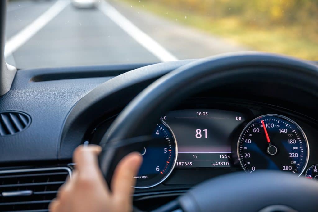 Visão do interior de um carro mostrando o painel de instrumentos, com o velocímetro marcando 81 km/h. Uma mão segura o volante enquanto o veículo está em movimento em uma estrada. Ao fundo, é possível ver a pista e a paisagem desfocada, indicando deslocamento.