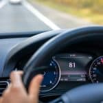 Visão do interior de um carro mostrando o painel de instrumentos, com o velocímetro marcando 81 km/h. Uma mão segura o volante enquanto o veículo está em movimento em uma estrada. Ao fundo, é possível ver a pista e a paisagem desfocada, indicando deslocamento.
