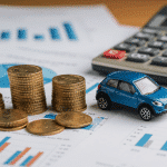 Um carro de brinquedo azul colocado em cima de documentos financeiros, com uma calculadora por perto. Existem moedas empilhadas, indicando um foco em finanças e orçamento. O fundo da imagem possui papéis com gráficos impressos.