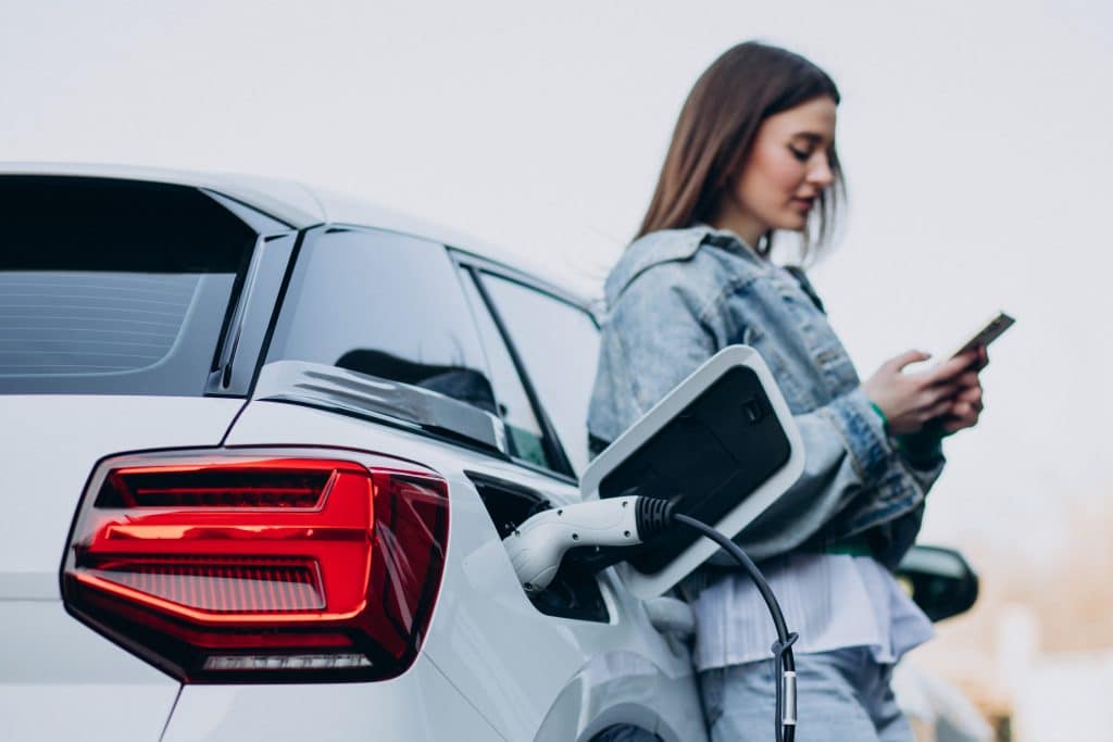 Uma mulher recarregando seu carro elétrico com uma estação de carregamento, enquanto usa seu smartphone. O veículo está conectado a um cabo de carregamento no porta de energia traseira, com o fundo desfocado sugerindo um ambiente urbano.