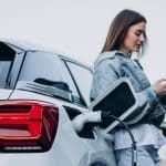 Uma mulher recarregando seu carro elétrico com uma estação de carregamento, enquanto usa seu smartphone. O veículo está conectado a um cabo de carregamento no porta de energia traseira, com o fundo desfocado sugerindo um ambiente urbano.