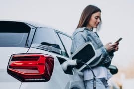 Uma mulher recarregando seu carro elétrico com uma estação de carregamento, enquanto usa seu smartphone. O veículo está conectado a um cabo de carregamento no porta de energia traseira, com o fundo desfocado sugerindo um ambiente urbano.