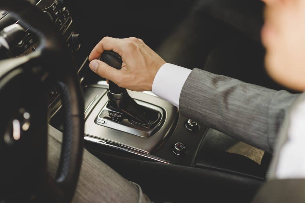 Mão de um homem de terno segurando a alavanca do câmbio automático de um carro. O painel central do veículo e o volante estão parcialmente visíveis, destacando o design sofisticado do interior do automóvel.