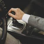 Mão de um homem de terno segurando a alavanca do câmbio automático de um carro. O painel central do veículo e o volante estão parcialmente visíveis, destacando o design sofisticado do interior do automóvel.