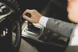 Mão de um homem de terno segurando a alavanca do câmbio automático de um carro. O painel central do veículo e o volante estão parcialmente visíveis, destacando o design sofisticado do interior do automóvel.