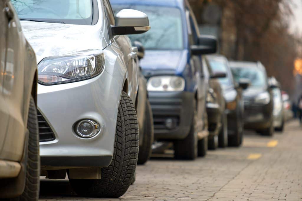 Imagem de um carro estacionado em uma rua com outros veículos ao fundo, mostrando detalhes dos pneus e parte da lateral.