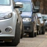 Imagem de um carro estacionado em uma rua com outros veículos ao fundo, mostrando detalhes dos pneus e parte da lateral.