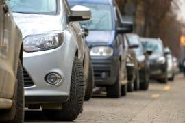 Imagem de um carro estacionado em uma rua com outros veículos ao fundo, mostrando detalhes dos pneus e parte da lateral.