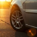 Imagem de um carro estacionado ao pôr do sol, com foco na roda dianteira e a estrada ao fundo.
