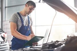 Mecânico vestindo macacão azul e segurando uma prancheta enquanto inspeciona o motor de um carro em uma oficina bem iluminada. O profissional faz anotações durante a verificação, garantindo a manutenção adequada do veículo
