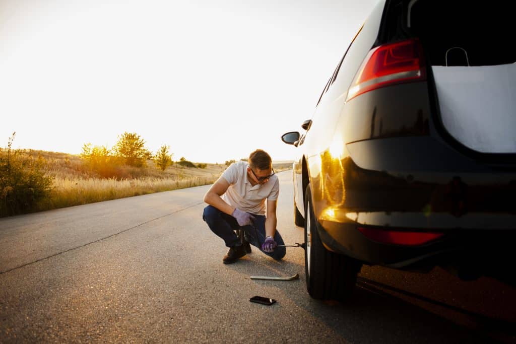 A imagem mostra um homem trocando o pneu de um carro na estrada durante o entardecer.
