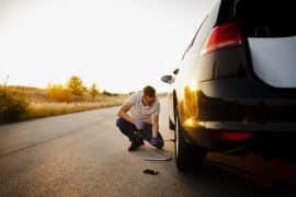 A imagem mostra um homem trocando o pneu de um carro na estrada durante o entardecer.